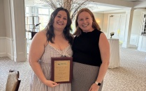 Zoom image: Two people stand together in a reception space, holding a framed award plaque from the University at Buffalo School of Law. A podium and light-filled windows appear behind them.