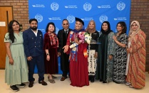 Zoom image: A group of people pose for a photo in front of a University at Buffalo School of Law backdrop. One person at the center wears graduation regalia and holds a bouquet of flowers, while others stand on either side in formal and traditional attire.