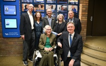 Zoom image: A group of attendees pose indoors in front of a wall display at the University at Buffalo School of Law. One person is seated in a wheelchair at the center, with others standing close behind.