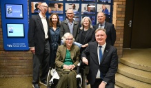 Zoom image: A group of attendees pose indoors in front of a wall display at the University at Buffalo School of Law. One person is seated in a wheelchair at the center, with others standing close behind.
