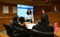 Zoom image: A small group of students sits around a table in a courtroom, writing on papers while two facilitators stand nearby, with a large screen behind them displaying a webpage titled &ldquo;Law Day.&rdquo;