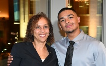Zoom image: Two people pose together indoors at a formal event, one holding a clear glass award, both wearing business attire, with illuminated city buildings visible through large windows behind them.