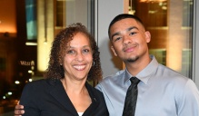 Zoom image: Two people pose together indoors at a formal event, one holding a clear glass award, both wearing business attire, with illuminated city buildings visible through large windows behind them.