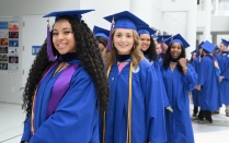 Zoom image: A group of graduates in blue caps and gowns walk indoors in a line. Tassels marked with &ldquo;26&rdquo; hang from the caps, and several graduates wear cords and stoles as they proceed through a bright hallway.