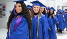 Zoom image: A group of graduates in blue caps and gowns walk indoors in a line. Tassels marked with &ldquo;26&rdquo; hang from the caps, and several graduates wear cords and stoles as they proceed through a bright hallway.