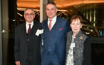 Zoom image: Three adults stand together indoors at a formal event, wearing suits and formal attire, with floor‑to‑ceiling windows behind them showing nighttime city lights.