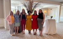 Zoom image: Six people stand side by side in a bright reception room, posing for a group photo in front of a tall decorative arrangement of branches with small lights. Cocktail tables and large windows are visible in the background.