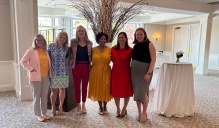 Zoom image: Six people stand side by side in a bright reception room, posing for a group photo in front of a tall decorative arrangement of branches with small lights. Cocktail tables and large windows are visible in the background.