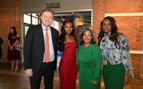 Zoom image: Four people stand together indoors at a formal event, dressed in business and evening attire. The setting features exposed brick walls, large windows, and soft decorative lighting. Other attendees and event signage are visible in the background, indicating a reception or social gathering space.