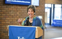 Zoom image: A person stands at a wooden podium with a University at Buffalo banner, holding a glass and speaking into a microphone. A brick wall and digital display screens are visible behind the podium.