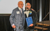 Zoom image: Two individuals in formal attire pose together on stage at an awards event, one holding a blue plaque, both wearing event name badges.