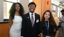 Zoom image: Three attendees wearing name badges pose together at an indoor networking event; one person in a light dress, one in a dark suit with a patterned tie, and one in a black top with a lanyard. Other guests and a refreshment table are visible in the background.