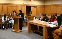 Zoom image: In a courtroom setting, a student stands at a wooden lectern reading from notes while other students sit at tables and benches nearby with papers and pens.