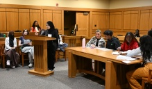 Zoom image: In a courtroom setting, a student stands at a wooden lectern reading from notes while other students sit at tables and benches nearby with papers and pens.