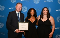 Zoom image: Three people stand in front of a University at Buffalo School of Law backdrop, posing for a photo. One person holds a framed certificate with the name &ldquo;Khalia J. Muir&rdquo; visible, while all three wear formal attire.