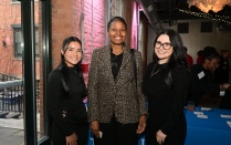 Zoom image: Three people stand together indoors at a formal event, wearing dark, professional attire. The setting includes exposed brick walls, large windows, and soft decorative lighting. A table with a blue tablecloth and event materials is visible behind them, indicating a reception or networking environment.