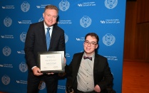 Zoom image: Two people pose in front of a University at Buffalo School of Law backdrop while holding a framed certificate. One person stands and holds the award, and the other is seated in a motorized wheelchair, both dressed in formal attire.