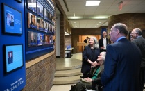 Zoom image: Several attendees gather in a hallway at the University at Buffalo School of Law, looking at a wall of digital and framed displays. A person seated in a wheelchair is positioned near the front, with others standing nearby.