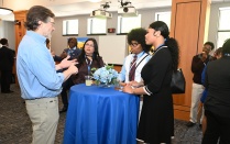 Zoom image: A small group stands around a tall table with a blue tablecloth and floral centerpiece while engaging in conversation at an indoor reception. Drinks and appetizers sit on the table, with additional attendees and screens visible in the room.