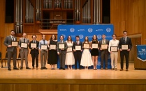 Zoom image: A group of students stands on a stage holding framed certificates, posing in front of a University at Buffalo School of Law backdrop. A large organ and wood-paneled walls are visible in the background.