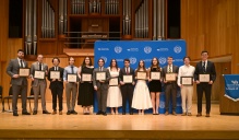 Zoom image: A group of students stands on a stage holding framed certificates, posing in front of a University at Buffalo School of Law backdrop. A large organ and wood-paneled walls are visible in the background.