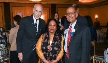 Zoom image: Three people in formal attire stand together at a banquet style awards event, with tables, chairs, and other attendees visible in the background.