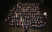 Zoom image: A large group of graduates wearing black caps and gowns are seated together in rows for a group photo in an auditorium. Many wear cords or stoles, and the dark stage setting surrounds the group.