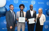 Zoom image: Four individuals pose in front of a University at Buffalo School of Law backdrop, with two holding framed Outstanding Oral Advocacy certificates during an awards presentation.