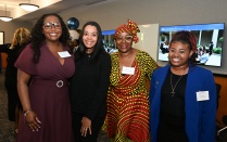 Zoom image: Four attendees wearing name badges stand side by side and pose for a photo at an indoor event; one person wears a patterned outfit with a matching headwrap, while others wear business or business-casual attire. A display screen and event d&eacute;cor appear in the background.