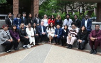 Zoom image: Group portrait of more than 20 individuals in business and professional attire seated and standing on tiered outdoor steps in a plaza surrounded by office buildings, trees, and landscaped greenery.