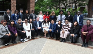 Zoom image: Group portrait of more than 20 individuals in business and professional attire seated and standing on tiered outdoor steps in a plaza surrounded by office buildings, trees, and landscaped greenery.