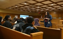 Zoom image: A presenter stands at the front of a courtroom-style room, speaking to a seated audience of students on wooden benches, with a large screen and an American flag visible behind the presenter.