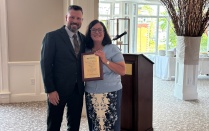 Zoom image: Two people pose together near a podium in an event room, with one holding a framed award plaque. A tall decorative branch arrangement and large windows are visible in the background.