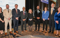Zoom image: Ten people in formal attire pose together at an indoor awards event, standing on a stage in front of a screen displaying names, with floral pins and event d&eacute;cor visible.