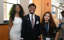 Zoom image: Three attendees wearing name badges pose together at an indoor networking event; one person in a light dress, one in a dark suit with a patterned tie, and one in a black top with a lanyard. Other guests and a refreshment table are visible in the background.