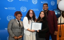 Zoom image: Four individuals pose in front of a University at Buffalo School of Law backdrop, with two holding a framed certificate of achievement during a recognition event.