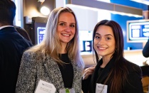 Zoom image: Two individuals standing together at an indoor networking event. Both are wearing name tags and professional attire. One person holds a drink with ice and a garnish. Digital displays and softly lit d&eacute;cor are visible in the background.