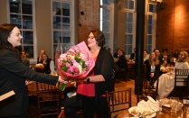 Zoom image: Woman smiling and presents a bouquet of flowers to another smiling woman. Seated attendees and banquet tables are visible in the background.