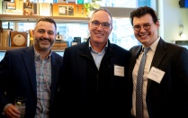 Zoom image: Three individuals standing closely together at an indoor event space. They are dressed in business or business casual attire and wearing name tags. Shelves in the background display books, decorative items, and small plants, with a window showing city buildings.