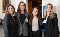 Zoom image: Four women standing indoors at a UB Law alumni event, smiling and posing together for a group photo. They are dressed in professional attire, with name badges visible and a University at Buffalo School of Law banner in the background.