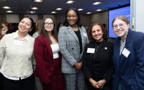 Zoom image: A group of people standing together and posing for a photo at an indoor event, dressed in business or business casual attire, with a presentation screen visible in the background.