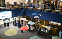 Zoom image: A large open area inside a library is set up for a career fair, with multiple employer tables arranged around the space. People in professional attire walk between tables and speak with representatives. Colorful umbrellas hang from the ceiling as decorations. The upper level of the library, visible in the background, has bookshelves and additional attendees walking along the balcony.
