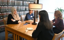 Zoom image: Four people sit around a wooden table in a library‑style room with shelves of law books covering the walls. A table lamp stands at the center of the table, which holds notebooks, a phone, and small dishes. The group appears engaged in conversation in a quiet indoor setting with natural light coming from a nearby window.