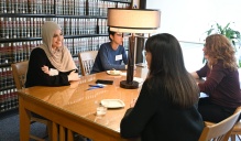 Zoom image: Four people sit around a wooden table in a library‑style room with shelves of law books covering the walls. A table lamp stands at the center of the table, which holds notebooks, a phone, and small dishes. The group appears engaged in conversation in a quiet indoor setting with natural light coming from a nearby window.