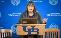 Zoom image: A person stands at a podium with the University at Buffalo logo, speaking in front of a blue UB School of Law backdrop with a laptop open.