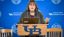 Zoom image: A person stands at a podium with the University at Buffalo logo, speaking in front of a blue UB School of Law backdrop with a laptop open.