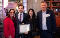 Zoom image: Four individuals standing together at an indoor event. One person in the center holds a framed certificate. All are wearing professional or business casual clothing and name tags. The background includes decorative lighting and shelving with various objects.