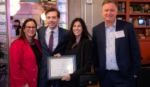 Zoom image: Four individuals standing together at an indoor event. One person in the center holds a framed certificate. All are wearing professional or business casual clothing and name tags. The background includes decorative lighting and shelving with various objects.