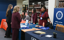 Zoom image: Representatives stand behind employer tables at a career fair inside a library, speaking with students who are visiting the booths. The tables are covered with branded tablecloths, informational materials, and small giveaways. Shelves of legal books line the background, and a banner for the Office of the New York State Attorney General is displayed on the right.