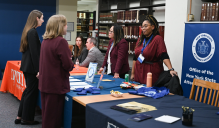 Zoom image: Representatives stand behind employer tables at a career fair inside a library, speaking with students who are visiting the booths. The tables are covered with branded tablecloths, informational materials, and small giveaways. Shelves of legal books line the background, and a banner for the Office of the New York State Attorney General is displayed on the right.